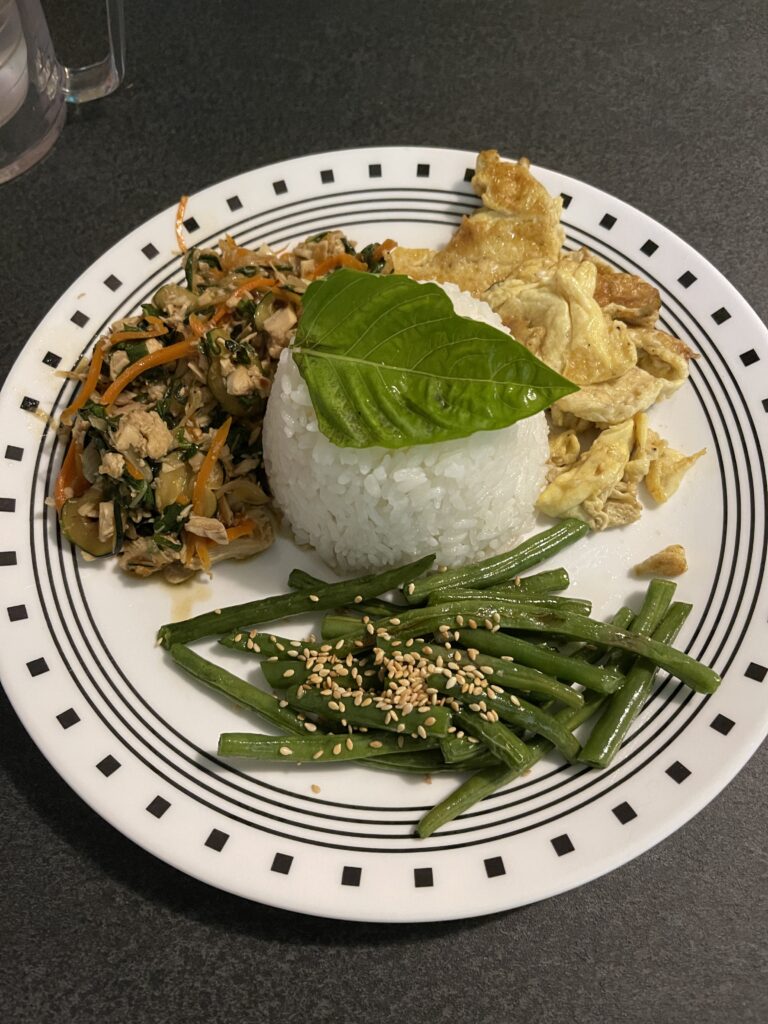 A white plate with black lines and squares around the edge. It has a molded pile of rice with a fresh basil leaf on top.  The remaining three foods are piled around the tidy rice. 