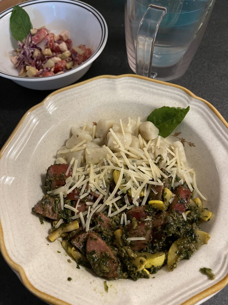 A round stoneware bowl contains gnocchi with pesto and sausage chunks and cheese. A bowl to the side contains veggies garnished with a basil leaf. 