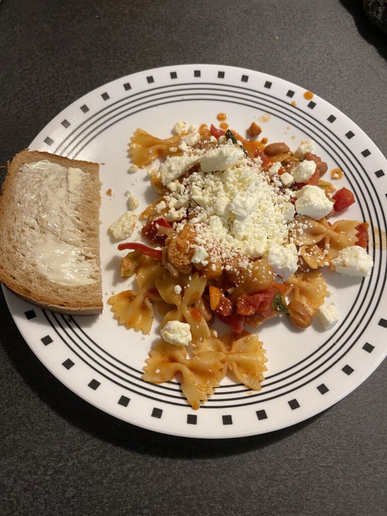 Tomato sauced bow tie pasta is heaped with veggies and feta cheese, with a side of buttered sourdough bread on a white plate with stripe and square black border. 