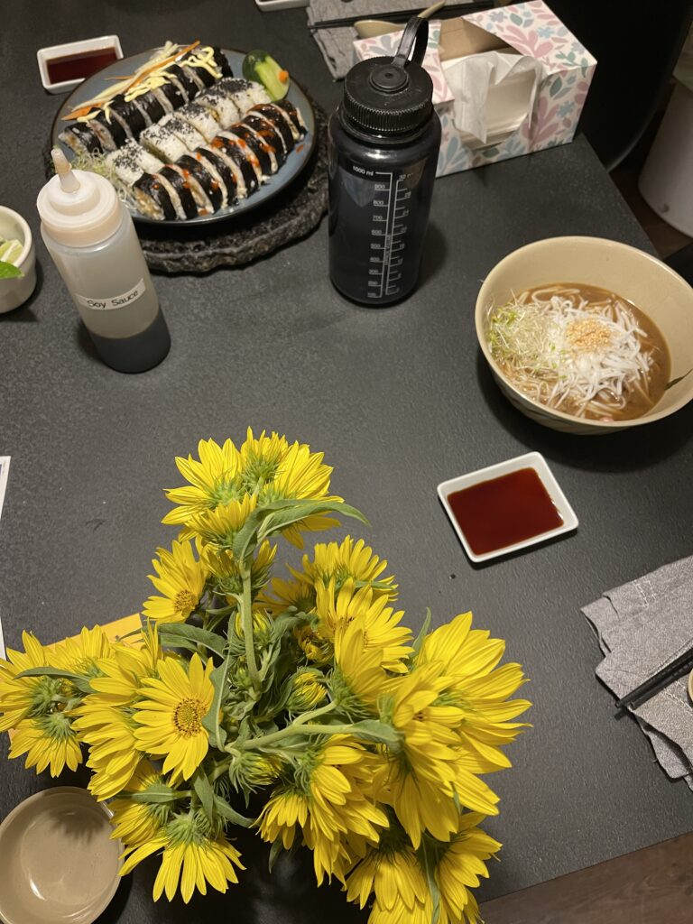 A bouquet of small sunflowers foregrounds a gray table with a noodle veggie soup and a plate with three rows of handmade sushi. 