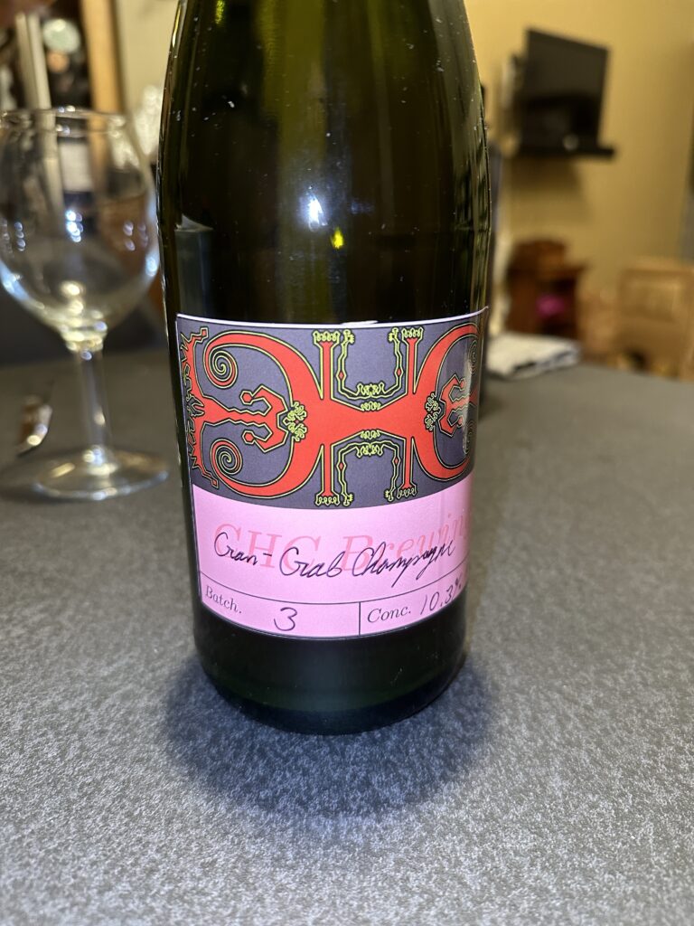 The champagne bottle (dark green glass) with the Chef’s hand designed reusable vinyl label.  The vintage area is pink with darker pink letters saying “CHC Brewing”. Handwritten notes specify “Cran + Crab Champagne, batch 3, 10.3% alcohol”. The logo is CHC with the first C reversed for symmetry. The letters are red with ornate, almost fractal, curlicues around them in a slate dark gray background. 