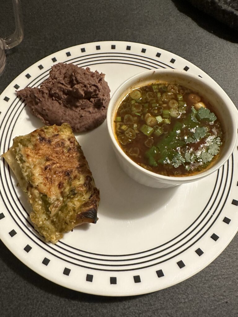 A ramekin of pozole, a pile of refried beans, and half a turkey enchilada with green sauce on a white plate edged with black lines and squares. 