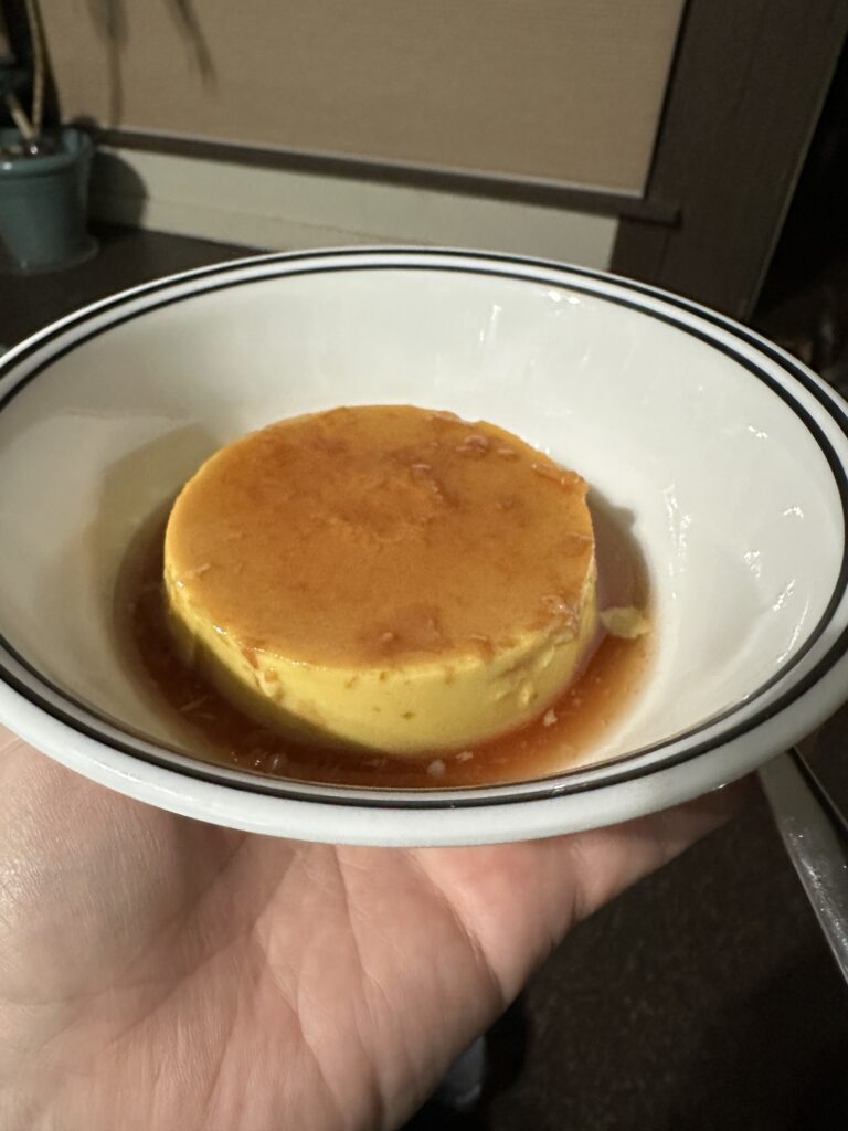 A perfect round egg-colored flan with an apple-tinted syrup in a white bowl with black stripe around the edge