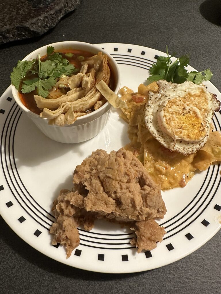 A ramekin of chicken tortilla soup garnished with cilantro, a side of refried beans, and chilaquiles topped with an egg. 